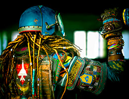 Person wearing colorful, Afrofuturistic flightsuit, with a view from the back. Dreadlocks, helmet and arm raised and hand holding cone at head level.