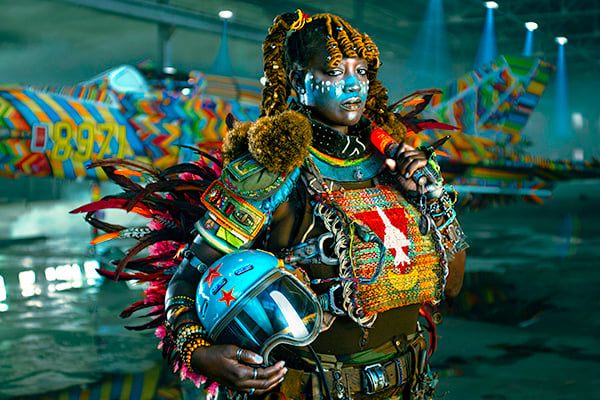 Black woman with a determined expression and wearing colorful regalia with beads, feathers, fabrics and holding blue pilot helmet, standing in front of a multicolored MiG-21 fighter jet.